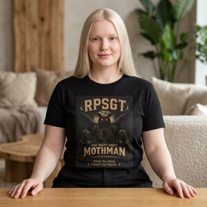 Könnte beinhalten: Ein schwarzes Kurzarm-T-Shirt mit einer Grafik eines geflügelten Wesens mit roten Augen. Der Text lautet: RPSGT, THE NIGHT SHIFT MOTHMAN, Guardian of Sleep Studies, While You Sleep I Watch The Signals. Das Design zeigt einen Mond im Hintergrund.