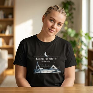 Peut inclure: Un t-shirt noir avec une impression graphique blanche représentant un croissant de lune, un compas, une équerre et un stylo sur un fond quadrillé. Le texte "Sleep Happens by Design." est imprimé en blanc au-dessus des outils de dessin.
