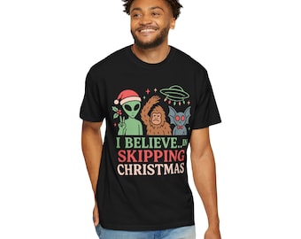 Op de afbeelding: Zwart t-shirt met een afbeelding van een alien met een kerstmuts, een Bigfoot en een vleermuisachtig wezen. De tekst "I Believe...in Skipping Christmas" staat onder de figuren gedrukt. Het ontwerp bevat een UFO en sterren.