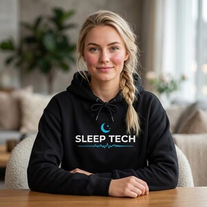 May include: Black hoodie featuring the white text "SLEEP TECH", a blue crescent moon, and a heartbeat line design. The hoodie has a drawstring and a front pocket. The model is wearing the hoodie.