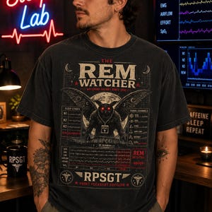 Könnte beinhalten: Ein anthrazitfarbenes T-Shirt mit einem Grafikdruck einer mottenartigen Kreatur mit leuchtend roten Augen. Das Design enthält den Text THE REM WATCHER, WATCHING DREAMS SINCE 1966, RPSGT und POINT PLEASANT DIVISION sowie verschiedene Schlafanalyse-Diagramme.