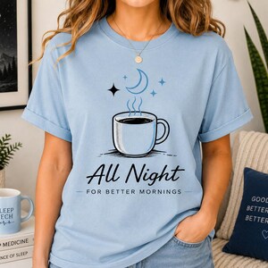 Peut inclure: Un t-shirt bleu clair à manches courtes orné d'un motif représentant une tasse de café avec de la vapeur, un croissant de lune et des étoiles. Le texte "All Night FOR BETTER MORNINGS" est inscrit en noir. Col rond et manches retroussées.
