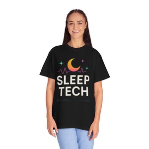 以下が含まれることがあります： 三日月、星、心拍線のグラフィックデザインが施された黒いTシャツ。グラフィックの下に「SLEEP TECH」というテキストが白で印刷されています。