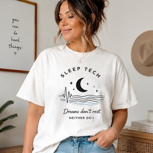 Könnte beinhalten: Ein weißes T-Shirt mit kurzen Ärmeln und einer schwarzen Grafik aus einer Mondsichel, Sternen und einer Herzschlaglinie über Wellenlinien. Der Text lautet SLEEP TECH, Dreams don't rest und NEITHER DO I in schwarzer Schrift.