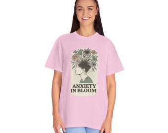 May include: A light pink t-shirt featuring a graphic of a person's profile with flowers blooming from their head. The text "ANXIETY IN BLOOM" is below the graphic. The shirt is made of a soft material.