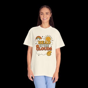 Op de afbeelding: Crèmekleurig T-shirt met de tekst "HEAD IN THE CLOUDS" in oranje en gele retro letters. Het ontwerp bevat een regenboog, een lachende zon, een bloem, wolken en een verfpalet. Het shirt is een casual stijl.