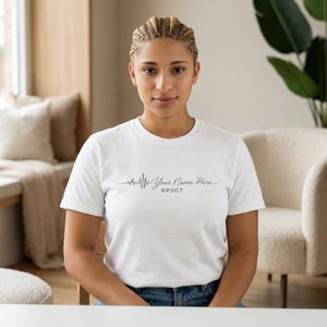 Peut inclure: T-shirt blanc avec un motif de ligne de battement de cœur noir et le texte "Your Name Here" et "RPSGT". Le t-shirt a un col rond et est fait d'une matière douce.