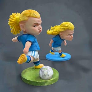 Peut inclure: Deux petites figurines de joueurs de football aux cheveux blonds attachés en queue de cheval, portant des maillots bleus et des shorts blancs. Une figurine est en mouvement de tir avec un ballon sur une base verte, l'autre sur une base bleue.