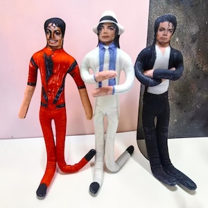 Puede incluir: Tres muñecos de tela planos que representan a Michael Jackson con diferentes atuendos. Uno lleva una chaqueta roja, otro un traje blanco con fedora y otro una chaqueta negra con camisa blanca. Cada muñeco tiene extremidades largas y finas.