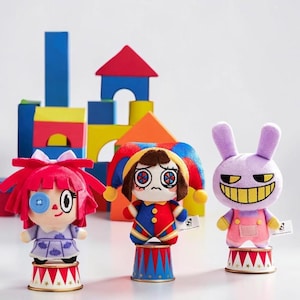 Puede incluir: Tres muñecos de peluche sobre pedestales individuales con temática de circo. El muñeco de la izquierda tiene pelo rojo y un ojo de botón azul. El central lleva un gorro de bufón rojo, azul y amarillo. El de la derecha es un conejo morado.