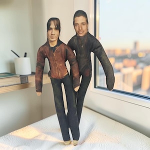 May include: Two custom fabric dolls featuring printed photographic portraits of individuals. One doll wears a brown jacket and dark pants, while the other wears a black jacket and dark pants. Both dolls have long, slender limbs and flat, stuffed bodies.