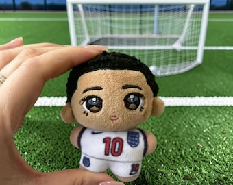 May include: A small plush doll with dark hair and large brown eyes, wearing a white sports jersey featuring the number 10 in red and a small blue emblem on the chest. The doll has a soft, rounded head and a compact body, designed as a collectible figure.