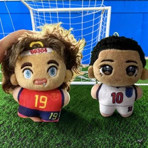 Op de afbeelding: Twee pluche poppen met voetbaltenues. De linkerpop heeft bruin haar, een witte hoofdband met de tekst 06304 en een rood shirt met het nummer 19. De rechterpop heeft zwart haar en een wit shirt met het nummer 10 en een blauw embleem.