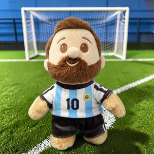 May include: A plush doll featuring a brown-haired figure with a beard, wearing a white and light blue striped soccer jersey with the number 10 printed on the front and an AFA logo, paired with black shorts, standing on a green field surface.