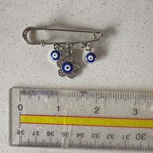 May include: A silver-toned safety pin with three dangling charms. Two charms are small blue and white evil eye beads, and the third is a flower-shaped charm with a central blue eye and clear crystals. The pin is approximately 1.5 inches long.