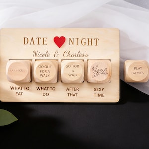 Può includere: Un gioco da tavolo in legno per una serata romantica con dadi. La tavola recita "Date Night" e "Nicole & Charles's". I dadi hanno opzioni come "Barbecue", "Fare una passeggiata" e "Sexy Time". C'è anche un cuore rosso sulla tavola.
