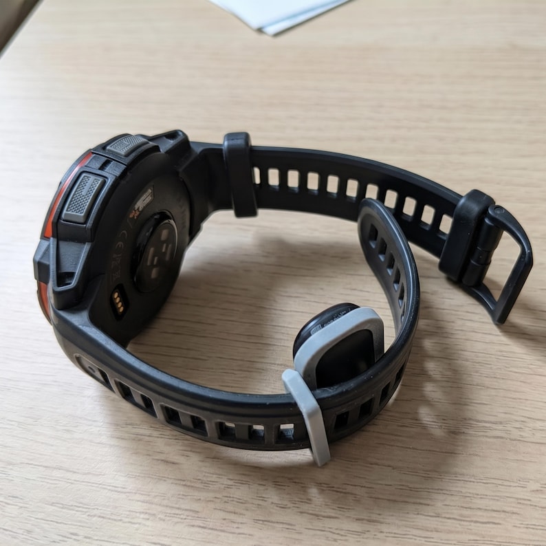 May include: A black digital sports watch with a textured silicone strap and a grey keeper loop. The watch case features a dark circular back with visible sensor components, charging contacts, and a prominent orange accent on the side edge.