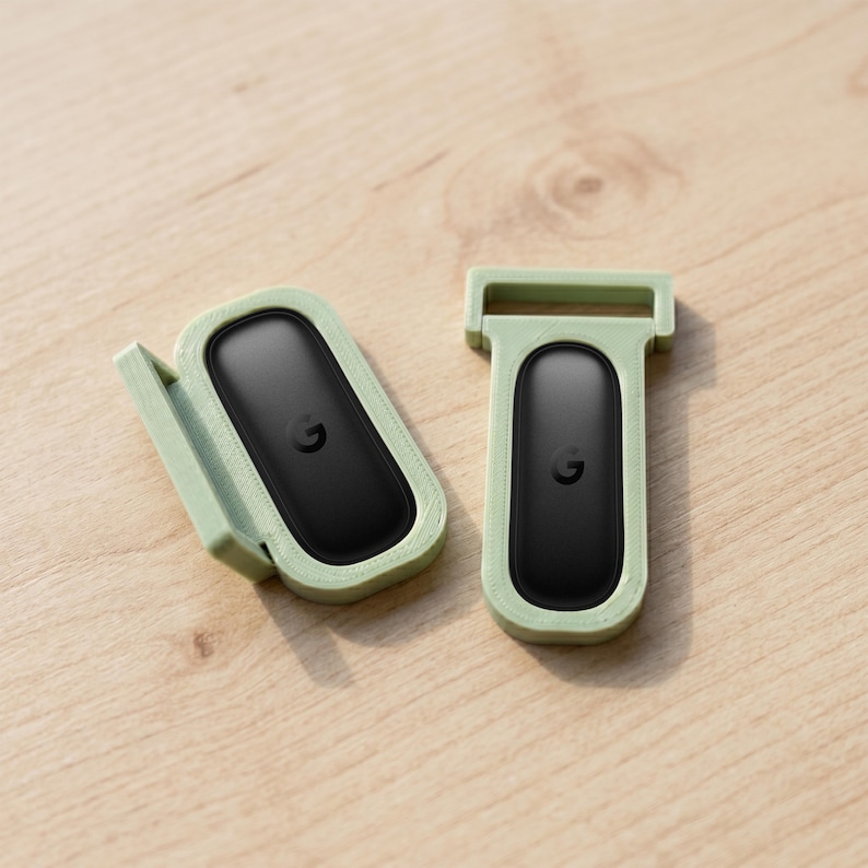 May include: Two black oval-shaped electronic trackers with a small letter G logo, each encased in a light sage green plastic mount. One mount features a side-hinged design, while the other has a rectangular loop at the top for attachment.