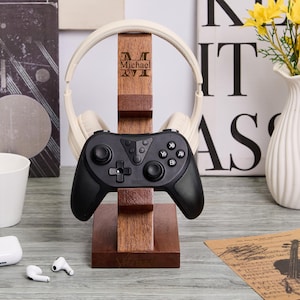 May include: A wooden headphone stand with a black game controller and cream-coloured headphones. The stand has a personalized name engraved on it. A white mug, wireless earbuds, and a vase of yellow flowers are also visible.