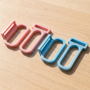 May include: Four rectangular plastic strap adjusters or sliders, featuring two in a soft pastel pink and two in a light sky blue. Each piece has a central open loop and a perpendicular bar at one end, designed for threading and securing fabric straps.