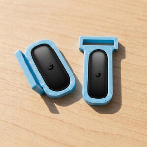 May include: Two light blue 3D-printed plastic holders, each containing a black oval-shaped electronic tracker with a small letter G logo centered on the surface. One holder is positioned horizontally with a side flap, while the other is vertical.