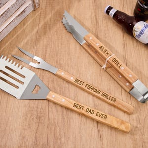 Puede incluir: Un juego de utensilios para barbacoa de acero inoxidable y madera, que incluye una espátula, un tenedor y unas pinzas. La espátula y las pinzas tienen el texto "BEST DAD EVER" y "ALEX' GRILL". También hay una botella de cerveza en la imagen.