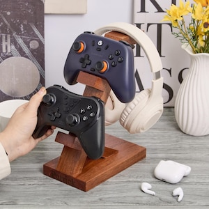 Puede incluir: Un soporte de madera para accesorios de juego con un mando de juego azul marino y otro negro, y unos auriculares color crema. También se ve un jarrón blanco con flores amarillas y un par de auriculares blancos.