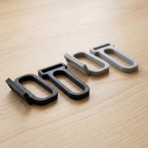 May include: Four small, rectangular, open-frame hooks arranged in a row. Two hooks are black and two are light gray. Each hook features a flat top bar, rounded sides, and a small angled tab extending from the base for hanging or securing items.