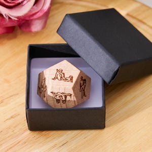 May include: A wooden, multi-sided die with etched figures, presented in an open black box. The die is light brown, and the figures are dark brown. The box sits on a wooden surface, with a pink rose in the background.