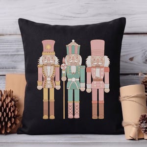 May include: A square black throw pillow featuring a centred graphic of three nutcracker figures. The nutcrackers are illustrated in pastel shades of pink, mint green, and yellow, each wearing a tall hat and uniform with detailed gold-toned accents.