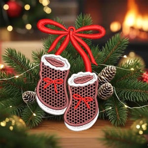 May include: Embroidered Christmas ornament featuring two small red and white baby booties with a red bow. The booties are surrounded by pine needles, pine cones, and a red bow at the top. The background includes a fireplace and Christmas lights.