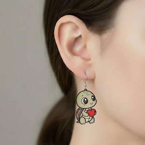 Peut inclure: Boucle d'oreille unique représentant une tortue de dessin animé tenant un cœur rouge. La tortue a une carapace verte avec un motif marron clair, de grands yeux et un visage souriant. La boucle d'oreille est suspendue à un crochet argenté.
