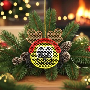 May include: A Christmas ornament featuring a cartoon dog face with reindeer antlers. The ornament is yellow, black, red, and white, with a honeycomb pattern. It hangs from a gold string and is surrounded by pine needles and pine cones.