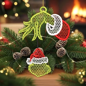 May include: A Christmas ornament featuring a green, red, and white Grinch design. The ornament is made of embroidered fabric and is hanging from a gold string. The ornament is surrounded by pine needles, pine cones, and fairy lights.