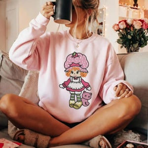May include: A light pink sweatshirt featuring a cartoon character with a strawberry hat, dress, and a cat. The character is in the center of the sweatshirt. The sweatshirt has long sleeves and a crew neck.