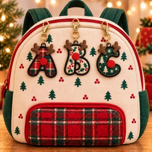 May include: A cream-colored backpack featuring a red and green plaid front pocket, green side panels, and a pattern of small green trees and red berries. Three gold-toned clips hang from the top, each holding a letter charm shaped like A, B, or C.