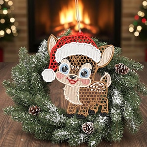 Könnte beinhalten: Ein Weihnachtsschmuck mit einem Cartoon-Hirsch, der einen rot-weißen Weihnachtsmannhut trägt. Der Hirsch hat braune Flecken und große blaue Augen. Er ist vor einem grünen Kranz mit Tannenzapfen platziert.