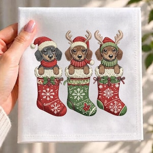 May include: A white fabric square featuring an embroidered design of three dachshunds inside Christmas stockings. The dogs wear festive hats and scarves, with two wearing reindeer antlers. The stockings are red and green with snowflake patterns.