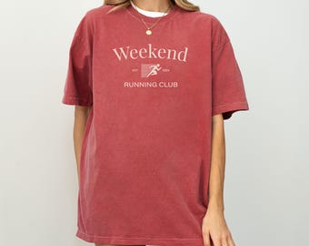 May include: A red-orange t-shirt with the words "Weekend Running Club" printed on the front. The text is in a light color, with a running figure graphic. The shirt has short sleeves and a crew neck.