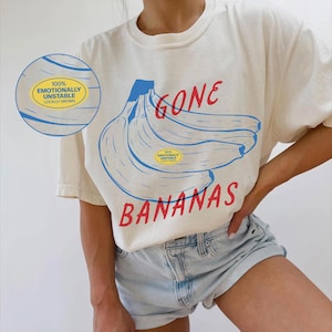 Peut inclure: Un t-shirt blanc avec un motif de banane bleue et le texte "Gone Bananas". Le t-shirt comporte également un autocollant jaune avec le texte "100% Emotionally Unstable Locally Grown".