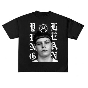 Puede incluir: Una camiseta negra de manga corta con un retrato en blanco y negro de una persona con corte de pelo tipo tazón. El texto YUNG LEAN está impreso en una fuente gótica blanca a ambos lados del retrato, con un pequeño icono de cara triste arriba.