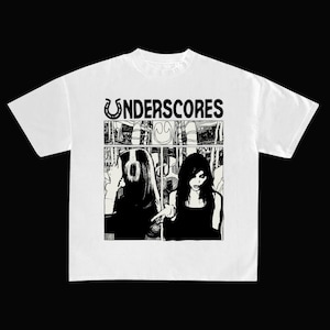 May include: A white short-sleeve t-shirt featuring a black graphic print on the chest. The design includes the text "UNDERSCORES" above a two-panel comic-style illustration showing a person with headphones and another person gesturing.
