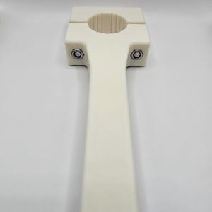 May include: A cream-colored, T-shaped clamp with a circular opening and a long handle. Two bolts secure the clamp's top section. The object is made of a smooth, solid material and is set against a plain white background.