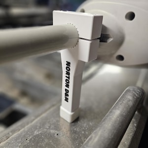 May include: A white, rectangular clamp with the text "NORTON D&M" in black, attached to a cylindrical pipe. The clamp is positioned against a larger white, rounded structure with several circular openings. The background is blurred.