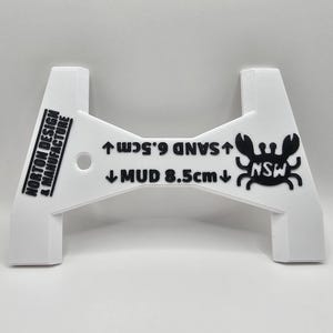 May include: White plastic tool with an A-frame design. The tool features black text: "NORTON DESIGN & MANUFACTURE", "SAND 6.5cm", and "MUD 8.5cm". A crab graphic and the letters "NSW" are also visible.