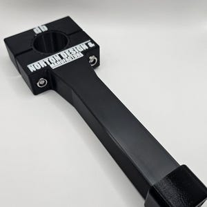 May include: Black metal clamp with a handle. The clamp has a square head with a circular opening and the text "NORTON DESIGN & MANUFACTURERS" in white. The handle is rectangular and ends with a square block.