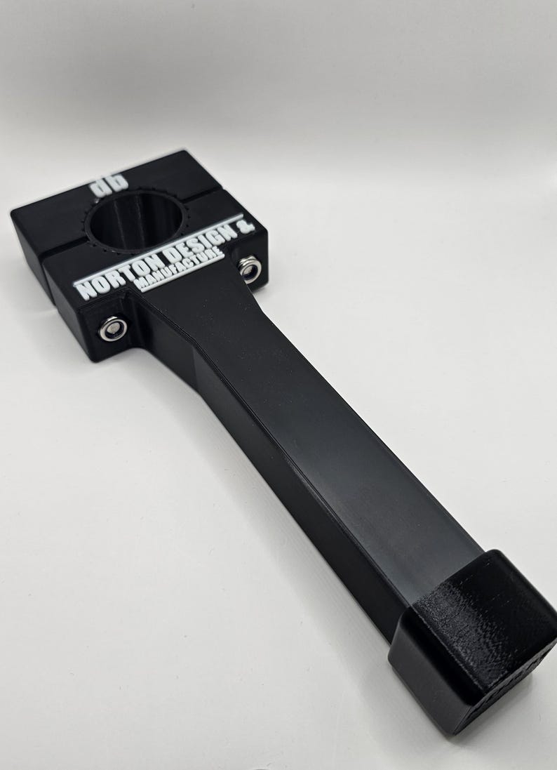 May include: Black metal clamp with a handle. The clamp has a square head with a circular opening and the text "NORTON DESIGN & MANUFACTURERS" in white. The handle is rectangular and ends with a square block.