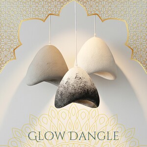 May include: Three pendant lights with textured, bell-shaped shades. One shade is black and white, the others are light grey and white. The lights hang from white cords against a white wall with gold accents. The text "GLOW DANGLE" is at the bottom.