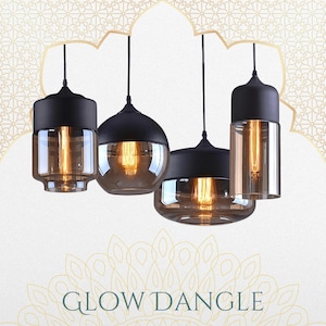 Op de afbeelding: Vier hanglampen met zwarte bovenkanten en heldere glazen kappen. Elke lamp heeft een warme, gloeiende gloeilamp binnenin. De lampen hangen aan zwarte snoeren tegen een witte achtergrond met een decoratief patroon. De tekst "GLOW DANGLE" staat onderaan.
