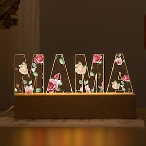 Könnte beinhalten: Beleuchtetes "MAMA"-Schild mit floralem Muster. Die Buchstaben sind mit rosa und weißen Rosen und grünen Blättern gefüllt, auf einem Holzsockel. Das Schild ist von innen beleuchtet und erzeugt ein warmes Leuchten.
